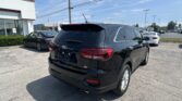 Black Kia SUV parked in a dealership lot with a glass storefront on the left and power lines overhead as a bright blue sky looms above.