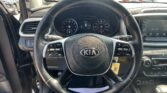 Inside view of a Kia car: steering wheel with Kia logo, instrument cluster, and center console with an illuminated infotainment screen; a parking lot is visible through the windows.