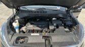 Car engine bay with hood raised; visible battery, coolant reservoir, and engine cover under a sunny parking lot roof.