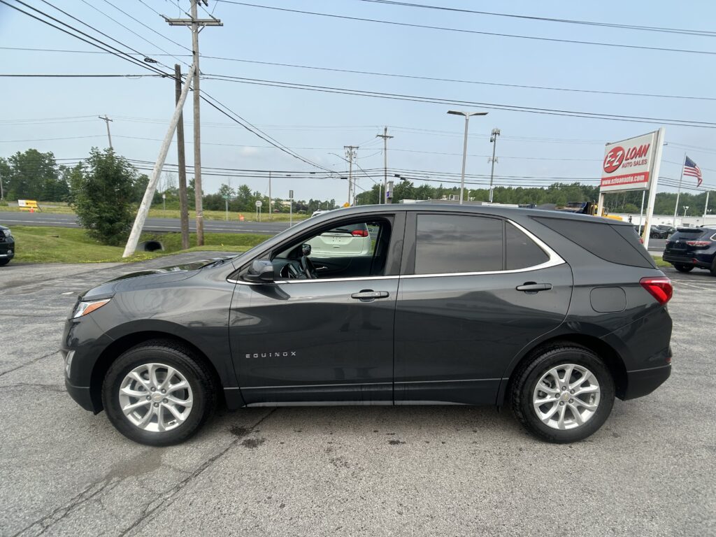 Gray Buick Enclave SUV parked in a dealership lot with an E-Z Loan Auto Sales sign and American flag in the background, power lines overhead.