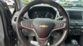Driver's view of the car interior showing the steering wheel with a Chevrolet emblem, dashboard gauges, and an in-dash infotainment screen, with a parking lot visible through the windshield.