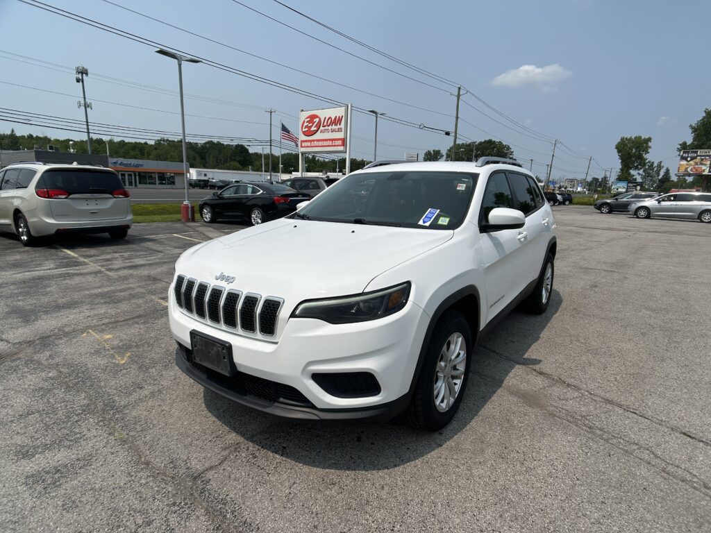 White Jeep SUV in a parking lot with other cars, power lines, and a red EZ LOAN sign in the background under a blue sky.