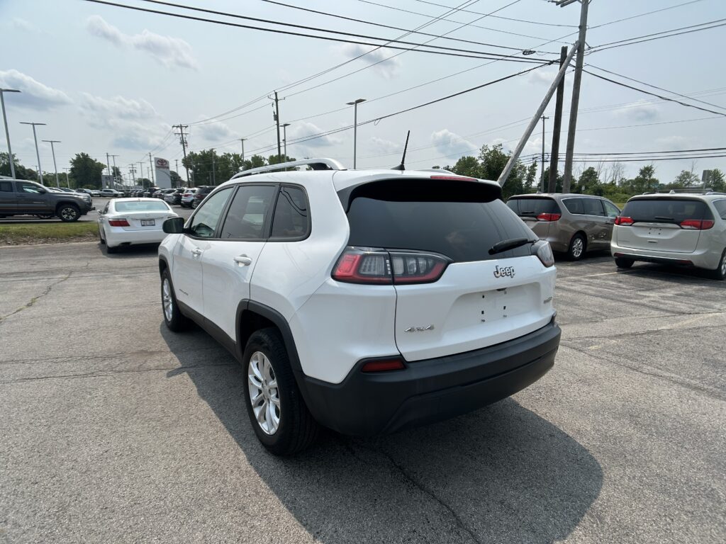 Rear three-quarter view of a white Jeep SUV in a crowded car dealership lot with power lines overhead.