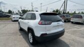 Rear three-quarter view of a white Jeep SUV in a crowded car dealership lot with power lines overhead.