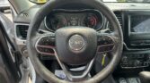 Steering wheel close-up inside a Jeep, with the instrument cluster and center touchscreen visible. Outside cars are in a parking lot.