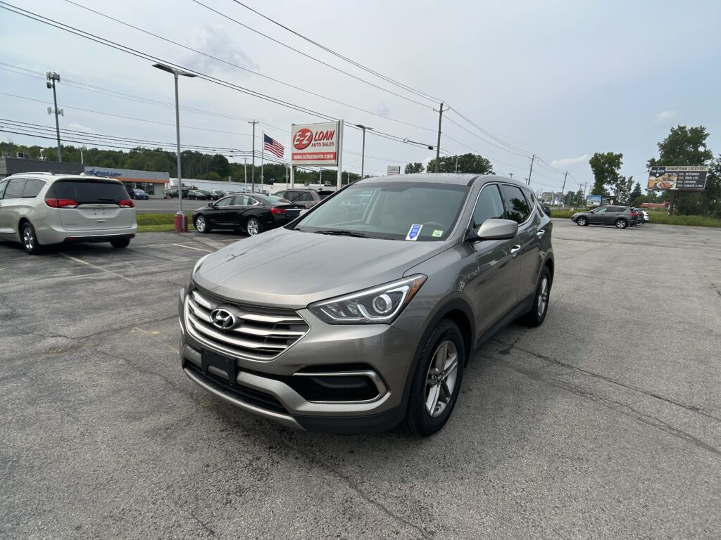 Silver Hyundai SUV in a car lot with an EZ Loan sign and American flag in the background, multiple parked cars around it.