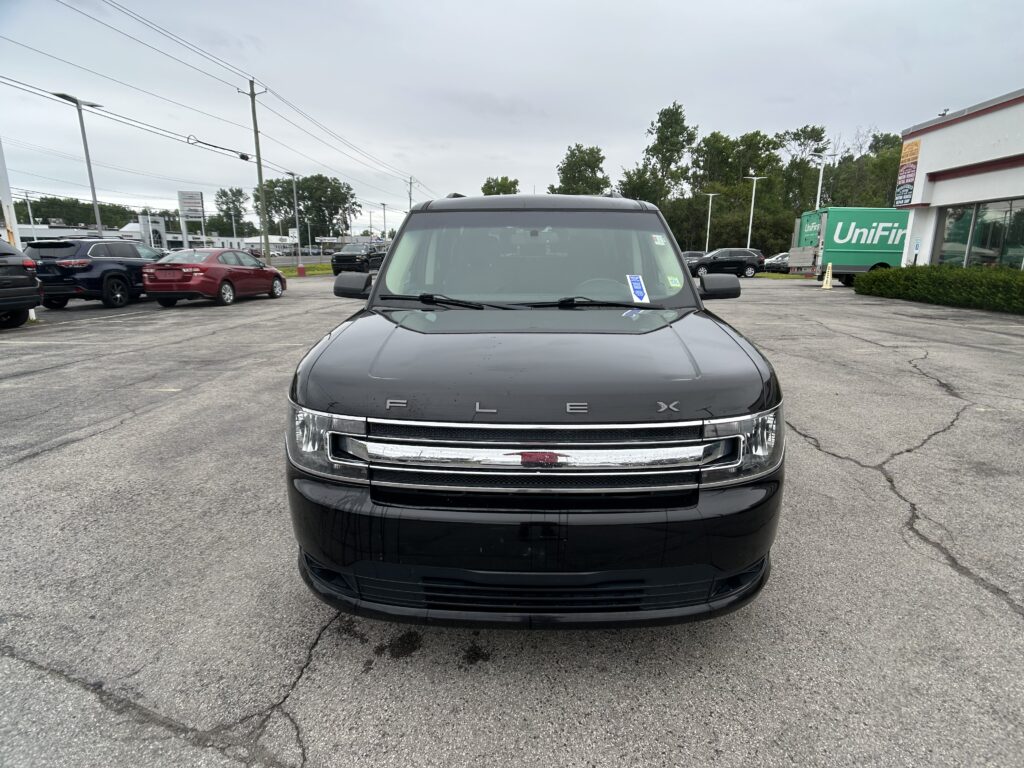Front view of a dark SUV parked in a lot, photo taken from the hood with other cars and a green UniFirst truck in the background. Fixes the scene's context for navigation.