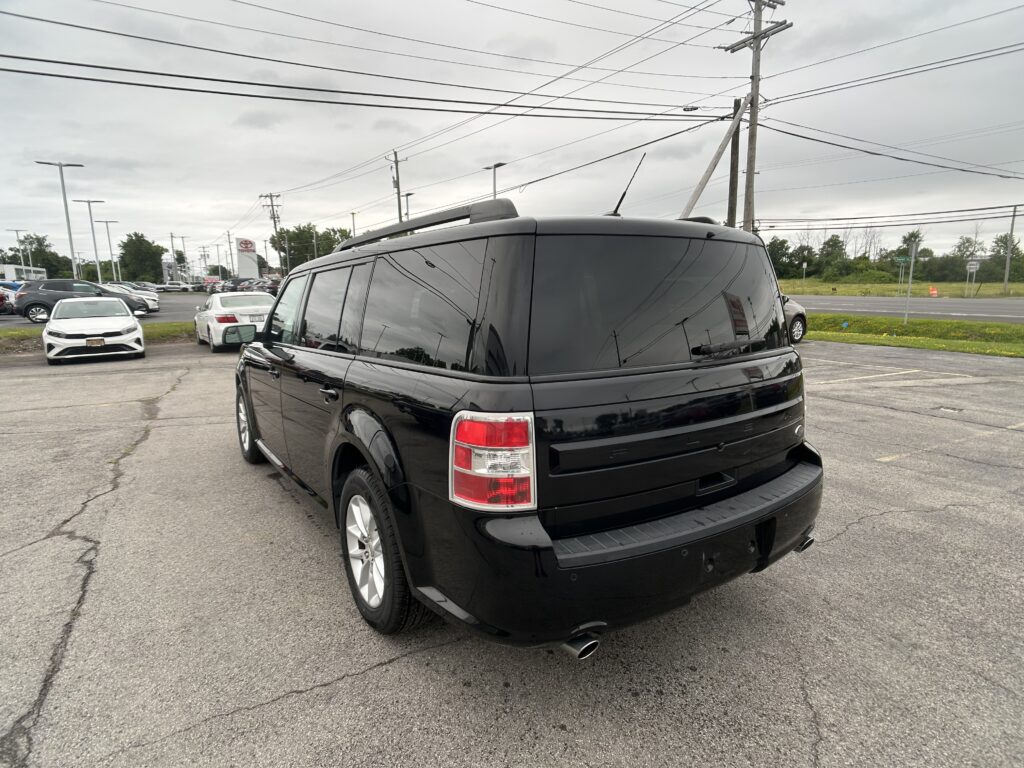 Rear view of a black SUV in a busy car dealership lot with power lines and cloudy sky above.