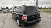 Rear view of a black SUV in a busy car dealership lot with power lines and cloudy sky above.