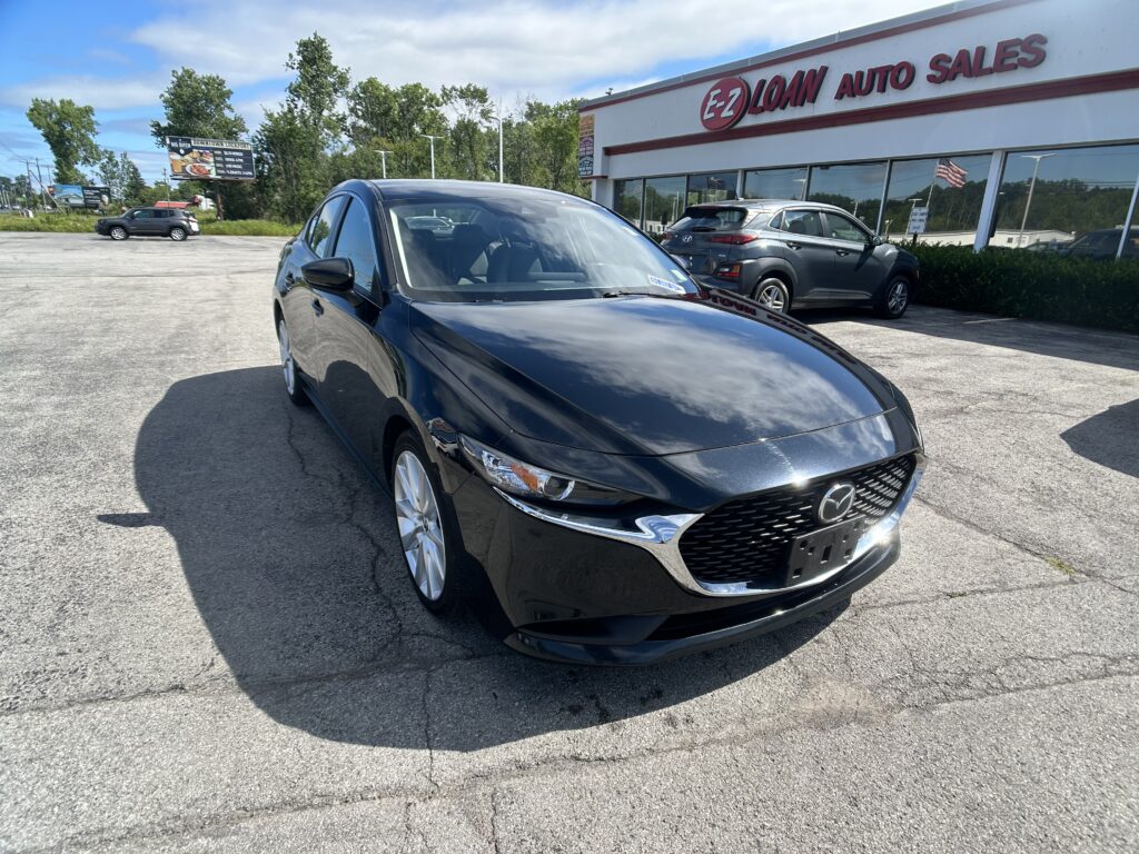 Black Mazda sedan in the foreground in a dealership lot with EZ Loan Auto Sales building in the background and a flag visible in the windows.
