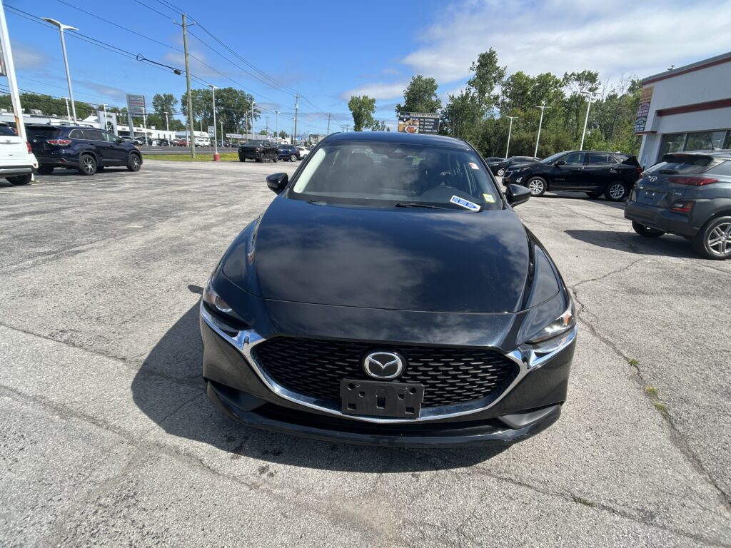Close-up of a dark gray Mazda front in a busy parking lot with other cars and a dealership building in the background on a sunny day.
