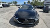Close-up of a dark gray Mazda front in a busy parking lot with other cars and a dealership building in the background on a sunny day.