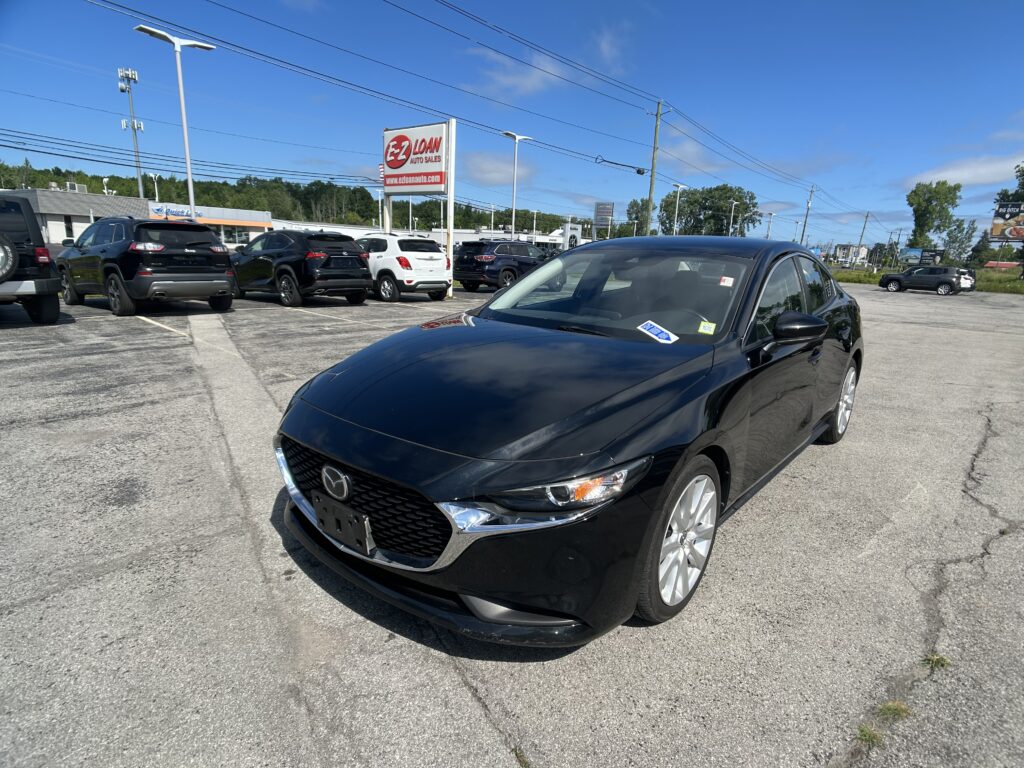 Close-up of a glossy black sedan in a car dealership lot with other vehicles and an EZ LOAN AUTO SALES sign in the background against a bright blue sky.