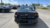 Rear view of a black Mazda3 in a busy car lot with other vehicles and a dealership in the background, under a blue sky.