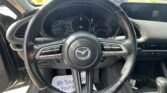 Close-up of a Mazda steering wheel from the driver’s seat, with the instrument cluster behind and a parking lot visible through the windshield.