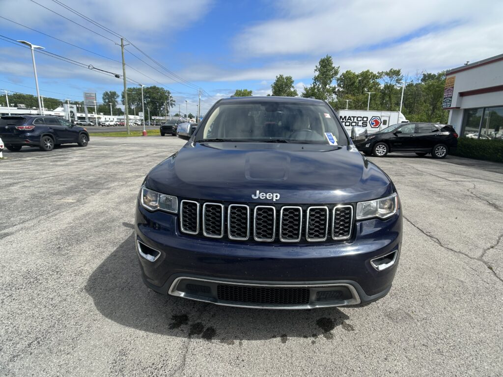 Front view of a dark blue Jeep SUV parked in a dealership lot with other vehicles and a building in the background in bright daylight.