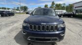 Front view of a dark blue Jeep SUV parked in a dealership lot with other vehicles and a building in the background in bright daylight.