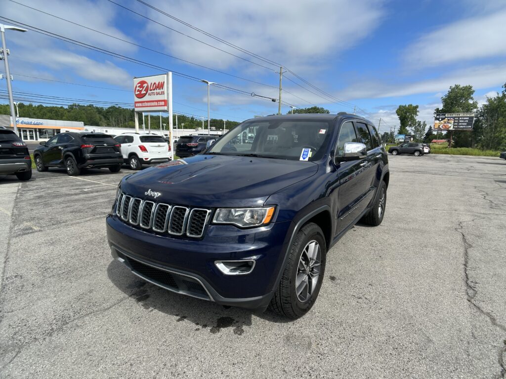 Dark blue Jeep SUV in foreground at a car dealership lot with several parked cars and a red EZ Loan Auto Sales sign in the background under a blue sky.