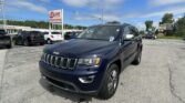 Dark blue Jeep SUV in foreground at a car dealership lot with several parked cars and a red EZ Loan Auto Sales sign in the background under a blue sky.