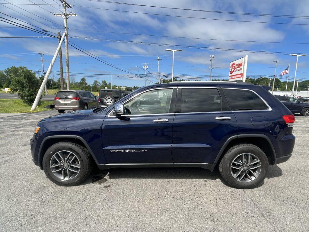 Blue Jeep Grand Cherokee parked in a dealership lot with overhead power lines and an EZ Loan Auto Sales sign in the background, American flag nearby.
