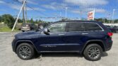 Blue Jeep Grand Cherokee parked in a dealership lot with overhead power lines and an EZ Loan Auto Sales sign in the background, American flag nearby.