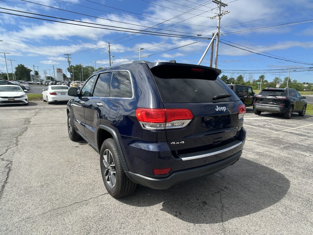 Rear three-quarter view of a dark blue Jeep SUV (4x4) in a sunny dealership parking lot with other cars nearby and power lines overhead, blue sky.