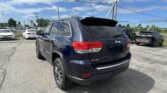 Rear three-quarter view of a dark blue Jeep SUV (4x4) in a sunny dealership parking lot with other cars nearby and power lines overhead, blue sky.