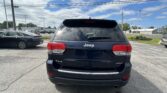 Rear view of a black Jeep SUV in a parking lot with other cars and utility poles in a bright, partly cloudy sky backdrop