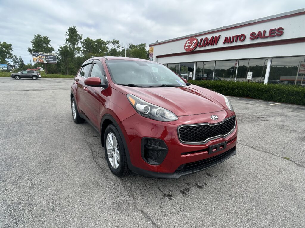 Red Kia SUV parked in the foreground outside a white building with 'EZ LOAN AUTO SALES' sign in red letters.