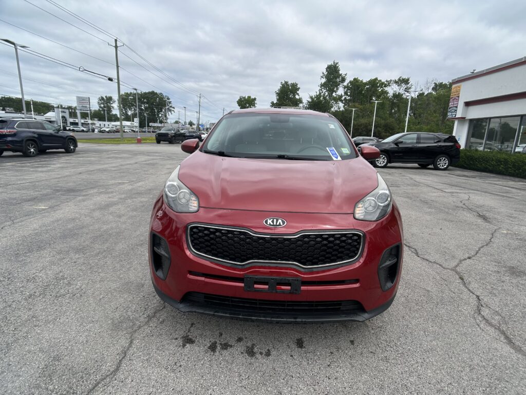 Front view of a red Kia SUV parked in a busy parking lot with a dealership building in the background.
