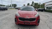 Front view of a red Kia SUV parked in a busy parking lot with a dealership building in the background.