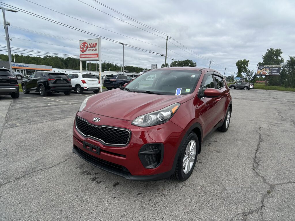 Front-right view of a red Kia SUV parked in a busy car lot under an overcast sky, with a large EZ LOAN auto sales sign in the background.