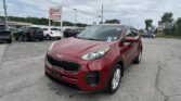 Front-right view of a red Kia SUV parked in a busy car lot under an overcast sky, with a large EZ LOAN auto sales sign in the background.
