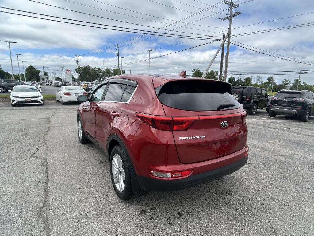 Red Kia Sportage rear in a packed dealership lot with other cars and tall utility poles overhead; cloudy sky above.