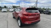 Red Kia Sportage rear in a packed dealership lot with other cars and tall utility poles overhead; cloudy sky above.