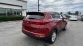 Rear three-quarter view of a red Kia SUV in a car dealership lot, with a white building and many cars under a cloudy sky.