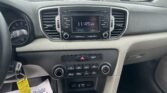 Center console of a car with a touchscreen display reading 11:45 AM; key fob hanging from the ignition on the left; papers on the passenger floor show 'We The People' text, visible through the windshield parking lot outside.