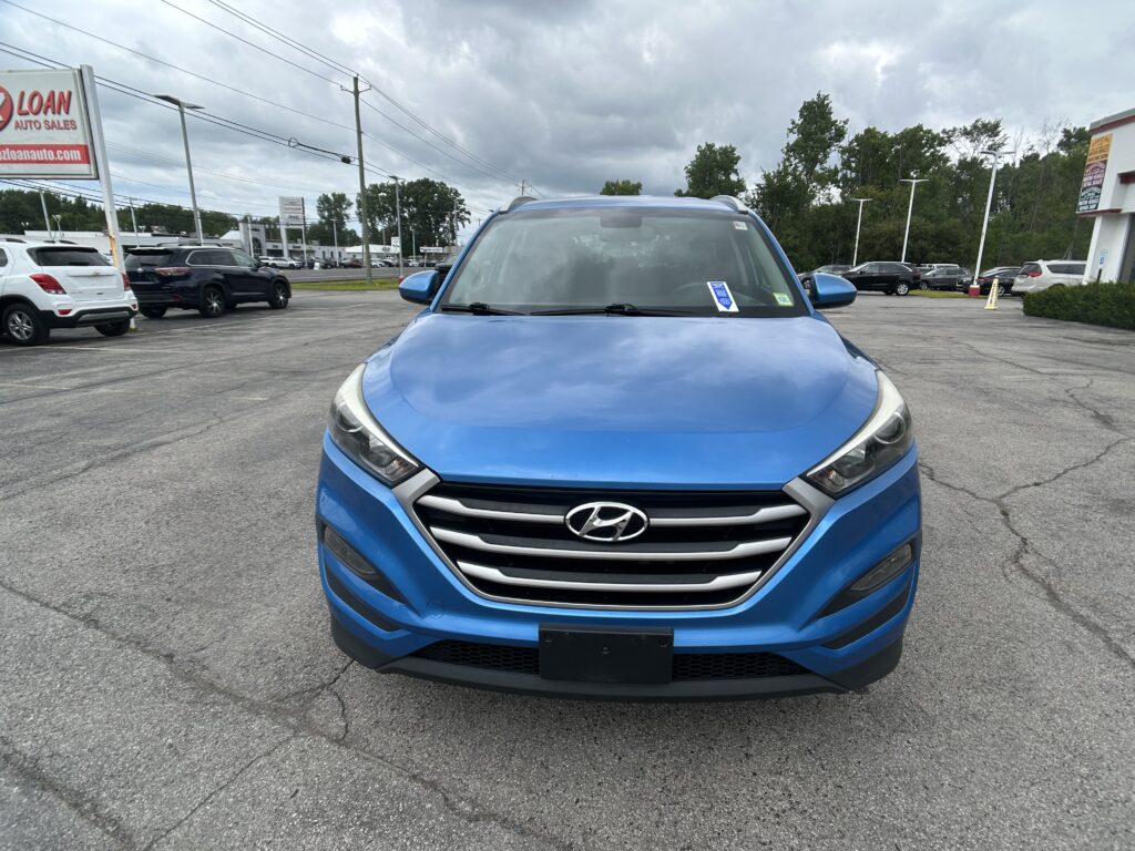 Blue Hyundai SUV photographed from the front in a car dealership lot with other vehicles and a cloudy sky overhead.