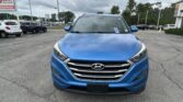 Blue Hyundai SUV photographed from the front in a car dealership lot with other vehicles and a cloudy sky overhead.