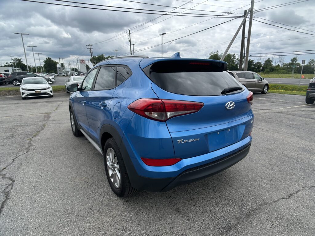 Blue Hyundai Tucson shown from the rear side in a car dealership parking lot with other cars and a cloudy sky above.