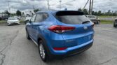 Blue Hyundai Tucson shown from the rear side in a car dealership parking lot with other cars and a cloudy sky above.