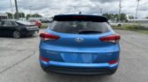 Rear view of a blue Hyundai Tucson SUV parked in a lot, with other cars and a cloudy sky above.