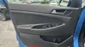Interior of a blue car door: black plastic panel with silver door handle and window control buttons; side mirror visible, parking lot scene outside through window