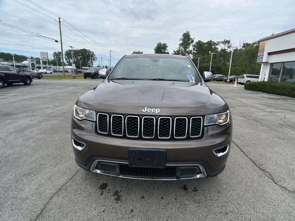 Front view of a brown Jeep SUV parked in a dealership lot with other cars and a building in the background.