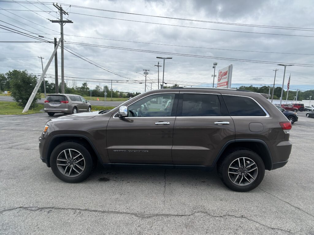 Brown Jeep Grand Cherokee in a dealership parking lot with power lines overhead and a cloudy sky behind it.