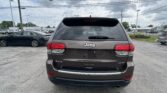 Rear view of a dark gray Jeep Grand Cherokee Limited 4x4 in a car dealership parking lot, with other vehicles and overcast sky above.