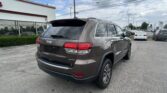 Brown Jeep Grand Cherokee Limited seen from rear three-quarter view in a dealership parking lot with other cars and a Toyota sign in the background.
