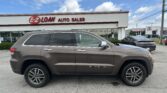 Brown Jeep Grand Cherokee SUV in the foreground at an auto dealership with an E-Z Loan Auto Sales sign on the building and other cars in the lot under a blue sky.