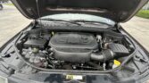 Open car hood revealing the engine bay with battery, hoses, and components in a parking lot.