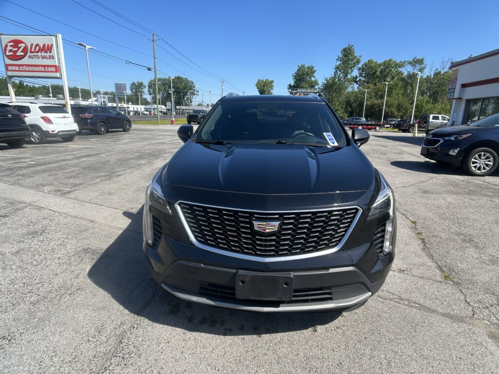 Front view of a dark blue Cadillac SUV in a car dealership lot with other vehicles and an EZ Loan sign in the background.